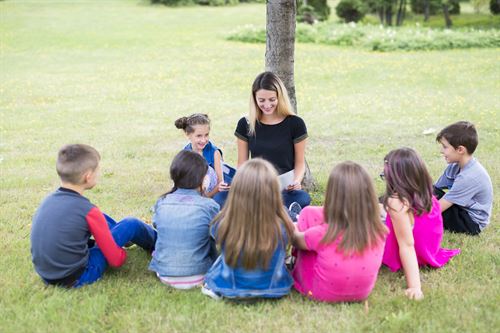A group of students sitting around a teacher reading to them under a tree